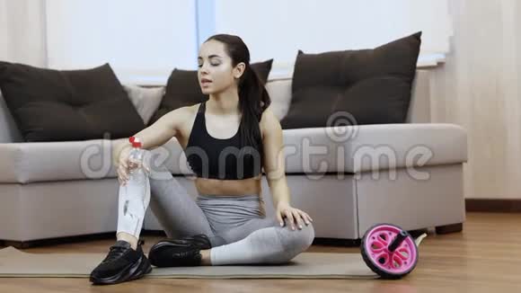 年轻女子在隔离期间在家锻炼坐在瑜伽垫上运动后喝水水化时间视频的预览图