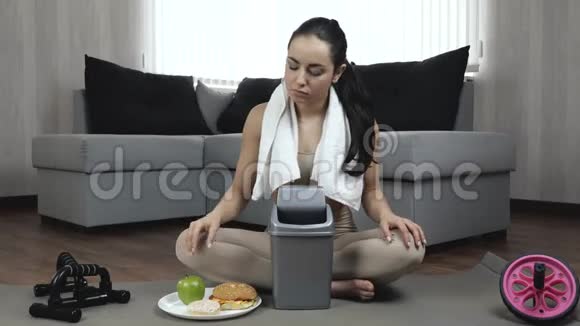 年轻女子在隔离期间在家锻炼坐在瑜伽垫上把汉堡和甜甜圈扔进垃圾桶带上你视频的预览图