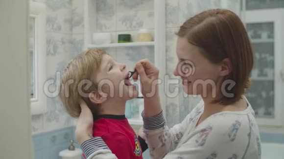 年轻的妈妈用牙线帮助穿红色睡衣的学龄前男孩刷牙有趣的家庭沐浴常规母亲视频的预览图
