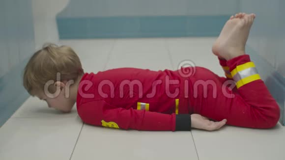 穿着消防员睡衣的金发学龄前男孩在浴室地板上玩得很开心在白地板上胡闹视频的预览图