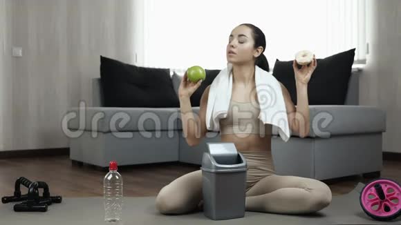 年轻女子在隔离期间在家锻炼吃绿色健康的苹果或者吃美味的油炸美甜甜圈之间的疑虑视频的预览图