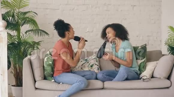 混合种族年轻有趣的女孩跳舞唱歌吹风机和梳子坐在沙发上姐妹享受休闲生活视频的预览图