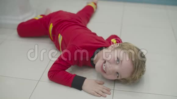 穿着消防员睡衣的金发学龄前男孩在浴室地板上玩得很开心孩子在白色地板上胡闹笑了视频的预览图