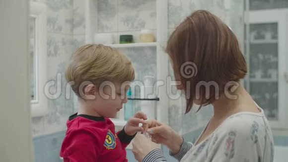 年轻的妈妈用牙线帮助穿红色睡衣的学龄前男孩刷牙有趣的家庭沐浴常规母亲视频的预览图