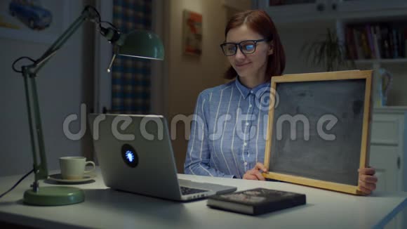 年轻微笑的30多岁的女人戴着眼镜拿着干净的空粉笔板看着家里的笔记本电脑在线教育程序视频的预览图