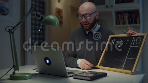 长着胡子的30多岁的男老师戴着眼镜在粉笔板上写数学用白色粉笔看着家里的笔记本电脑网上在线视频的预览图