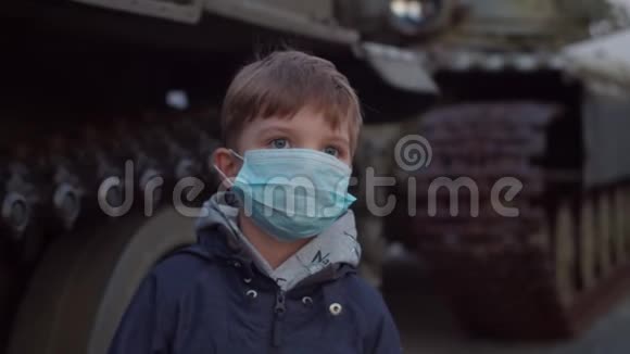 在冠状病毒COVID19大流行爆发期间戴医用防护面罩的男孩在军用机器旁站立并观看视频的预览图