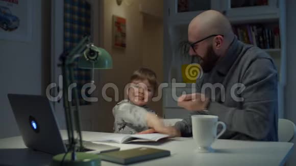 年轻的30岁的父亲在家里做网上作业时给儿子5分网上学习的胡子男和小男孩视频的预览图
