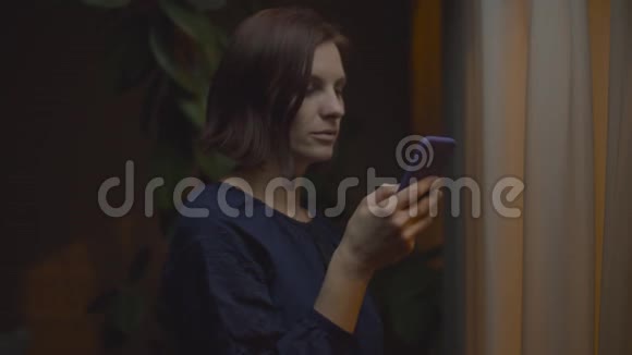 30多岁的女性穿着民族衬衫站在晚间灯光窗口在黑暗的房间里使用智能手机视频的预览图
