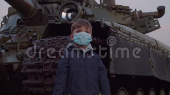 戴医用防护面罩的男孩在19日冠状病毒大流行爆发时站在军用机器旁观看视频的预览图