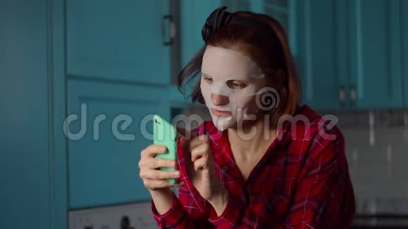 年轻的30多岁的女人用手机在脸上戴着白色的化妆面具在家里做自拍的鬼脸女人美丽视频的预览图