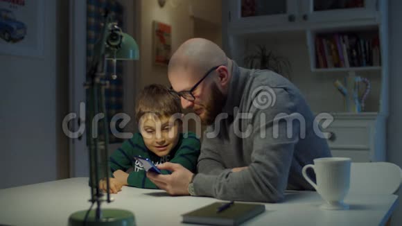 年轻的30岁父亲和上学的儿子在家看手机戴眼镜的大胡子还有带手机的男生视频的预览图