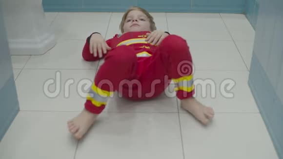 穿着消防员睡衣的金发学龄前男孩在浴室地板上玩得很开心在白地板上胡闹视频的预览图