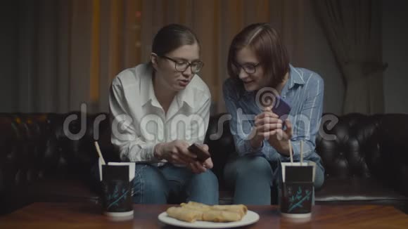 两个戴着智能手机眼镜的女性朋友在深夜坐在沙发上微笑和交谈亚洲盒装食品视频的预览图