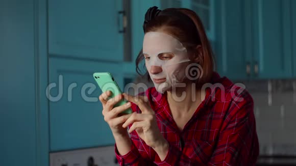 30多岁的年轻女性在家里使用手机脸上戴着白色化妆面具蓝色厨房的女人美容程序视频的预览图