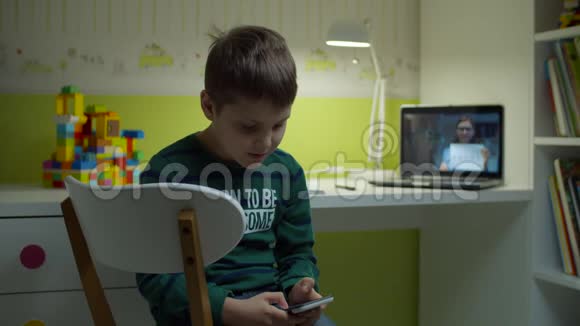 无聊的男生坐在书桌前拿着手机一边和女老师在笔记本电脑屏幕上在线上课视频的预览图