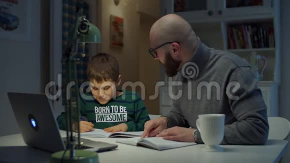 年轻的30岁的父亲和上学的儿子在家做网上作业戴眼镜的胡子男在网上学习视频的预览图