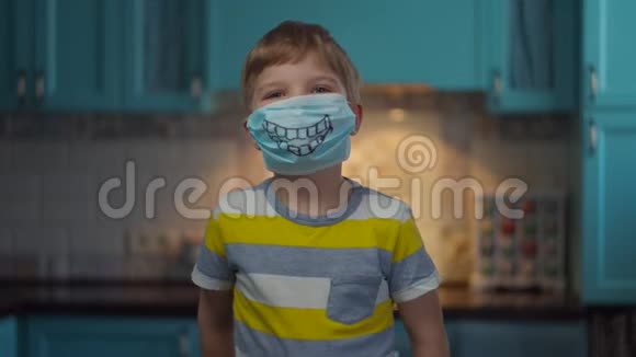 戴着医疗面具的男孩微笑着看着相机的肖像带着滑稽医疗面具的孩子在家中与家人隔离视频的预览图