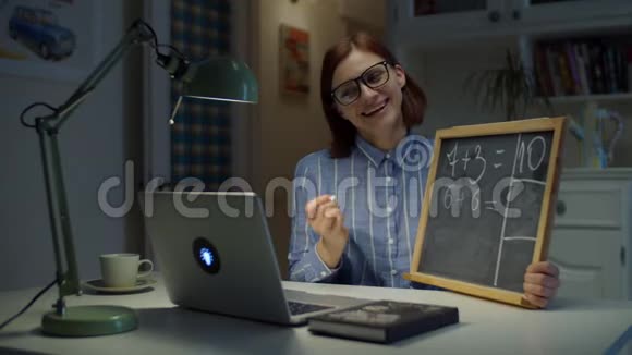 30岁的眼镜女老师用白色粉笔在黑板上书写简单的数学方程式看着笔记本电脑视频的预览图