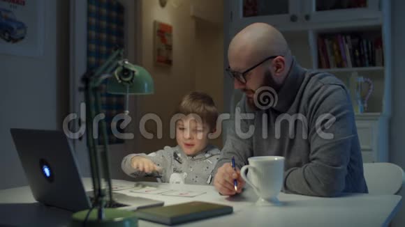 年轻的30岁的父亲和学龄前儿童在家做在线家庭作业小男孩出示卡片正确回答笔记本电脑视频的预览图