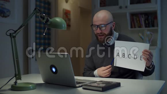 30多岁的留着胡子的戴眼镜的男人拿着一张写着字母表的纸看着家里的笔记本电脑在线教育视频的预览图