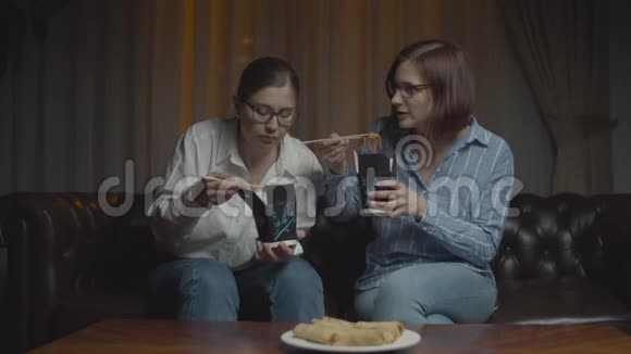 两个戴眼镜的女朋友用筷子吃亚洲盒饭女人会在深夜的沙发上搞笑聊天视频的预览图