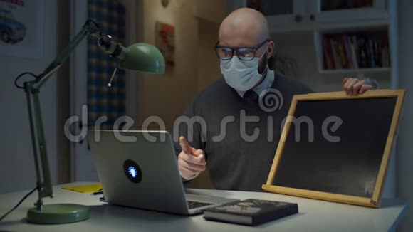 30多岁的大胡子男戴着医用口罩脸上写着COVID19黑板上写着白色粉笔看着家里的笔记本电脑视频的预览图
