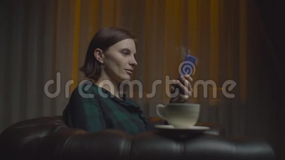30岁的女性成人微笑着使用智能手机深夜坐在舒适的扶手椅上喝咖啡并配有路灯视频的预览图