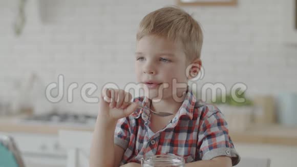 金发小男孩在白色厨房里慢悠悠地吃着玉米片和纯素牛奶看着平板电脑视频的预览图