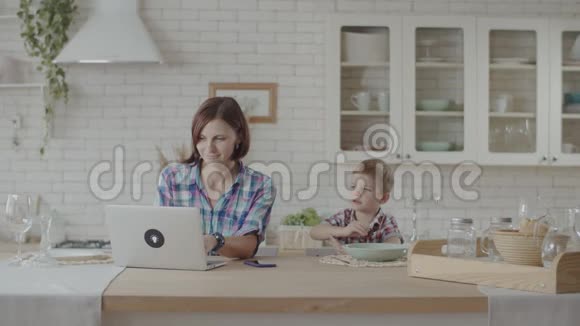 年轻的30岁的母亲和儿子坐在厨房里一边看小玩意一边吃零食快乐的家人一起吃饭视频的预览图