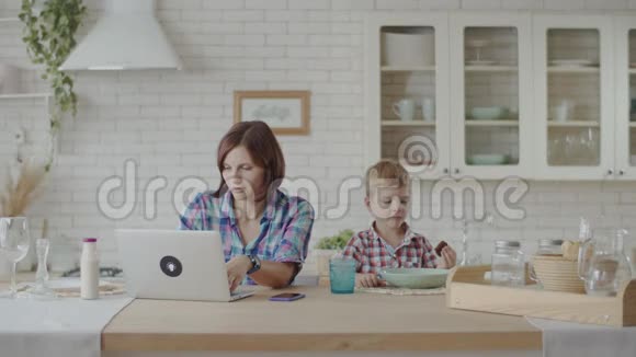 年轻的30岁的母亲和儿子坐在厨房里一边看小玩意一边吃零食快乐的家人一起吃饭视频的预览图