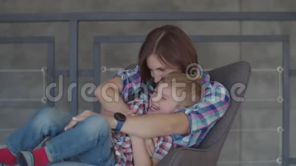 年轻的30多岁的黑发母亲和金发蹒跚学步的儿子坐在扶手椅上玩快乐的女人和男孩慢慢地笑视频的预览图