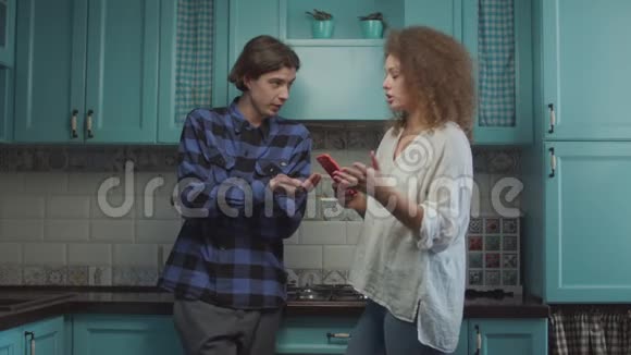 20多岁的年轻夫妇站在家里的蓝色厨房里吵架女朋友拿着手机大声指责视频的预览图