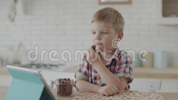 可爱的金发小男孩吃着巧克力球在白色厨房里慢悠悠地看着平板电脑视频的预览图