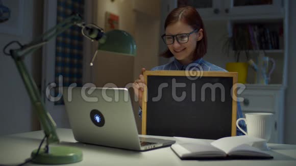 30多岁的年轻女性戴着眼镜手里拿着干净的空粉笔板看着家里的笔记本电脑在线教育过程工作视频的预览图
