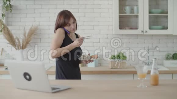 年轻的黑发女人穿着黑色连衣裙一边在明亮的厨房里散步一边吃寿司欧洲女性吃日本食物视频的预览图