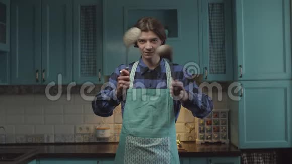 20多岁的年轻黑发男子穿着围裙手里拿着厨房用具站在蓝色的厨房里用滑稽的方式移动双手视频的预览图