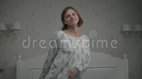 穿着睡衣的30多岁的年轻女子在床上用无线耳机欣赏音乐闭眼跳舞的女性视频的预览图