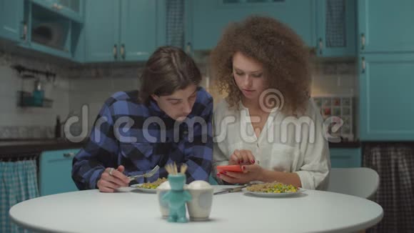 一对20多岁的年轻夫妇在蓝色的厨房里用小玩意儿吃饭男人和女人用手看手机视频的预览图