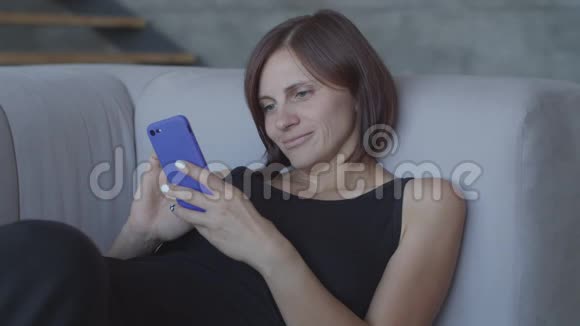 漂亮的30多岁的女人喜欢坐在舒适的沙发上用智能手机聊天穿着黑色裙子的漂亮女性视频的预览图