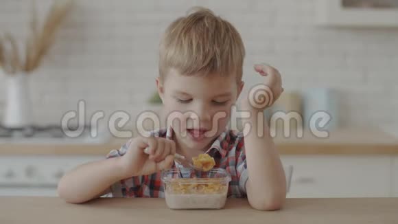 金发的蹒跚学步的男孩吃玉米片与纯素牛奶和喝苹果汁在白色厨房慢动作视频的预览图
