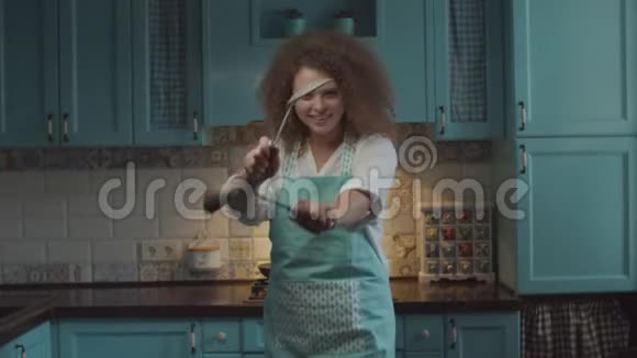 年轻卷发20多岁围裙女人手拿厨具站在蓝色厨房里用搞笑的方式移动双手视频的预览图