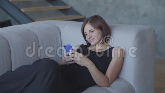 漂亮的30多岁的女人喜欢坐在舒适的沙发上用智能手机聊天穿着黑色裙子的漂亮女性视频的预览图