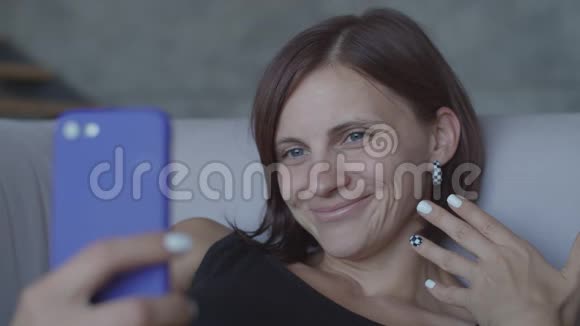 漂亮的30多岁的女人喜欢坐在舒适的沙发上用智能手机聊天穿着黑色衣服的漂亮女人视频的预览图