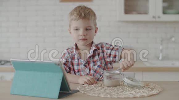 金发小男孩在白色厨房里慢悠悠地吃着玉米片和纯素牛奶看着平板电脑视频的预览图