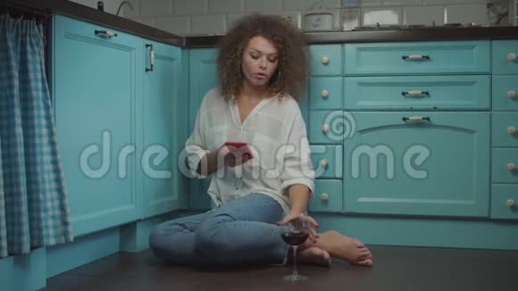 年轻的卷发20多岁的女人手里拿着手机喝着一杯红酒坐在厨房的地板上女性视频的预览图