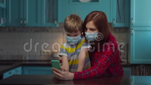 30多岁的母亲和男孩戴着医疗面具用手机与亲戚们在线交谈挥手致意家庭视频的预览图
