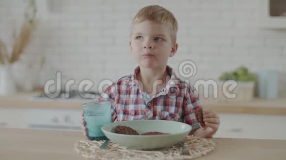 可爱的金发幼儿男孩喝酸奶和吃巧克力饼干在更白色的厨房慢动作视频的预览图
