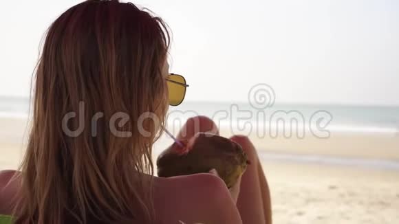一个年轻的女人在大海的背景下轻松地喝椰子视频的预览图