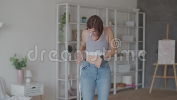 年轻女子在卧室里穿着旧牛仔裤欣赏减肥的结果微笑的女人测量她的腰视频的预览图
